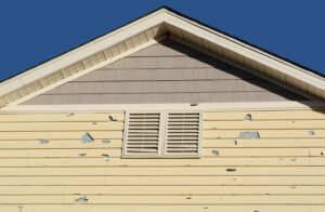 Yellow horizontal lap siding with multiple areas of peeling and flaking paint exposing the underlying gray surface, beneath a gable with shingle siding and white attic vents