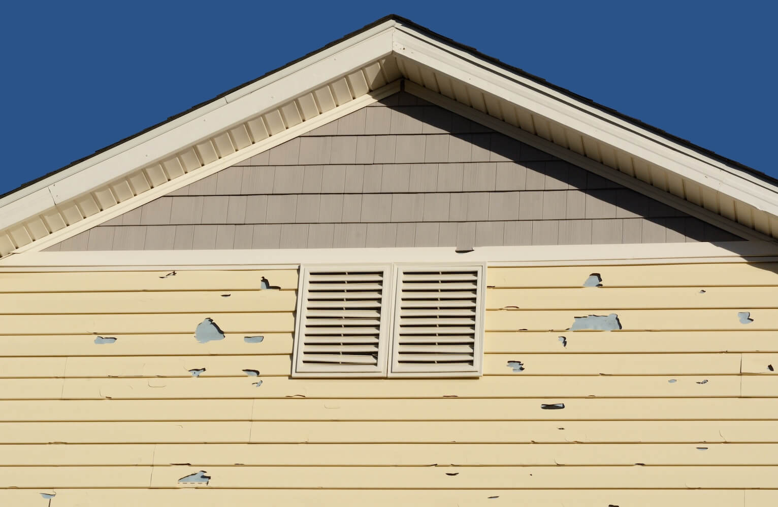 Yellow horizontal lap siding with multiple areas of peeling and flaking paint exposing the underlying gray surface, beneath a gable with shingle siding and white attic vents
