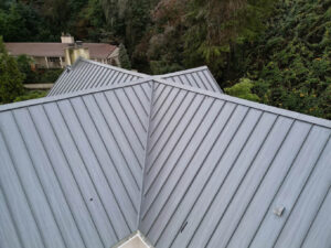 Aerial view of a silver gray standing seam metal roof with multiple intersecting roof slopes, raised panel seams, and metal hip and valley flashing